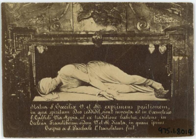 Black and white photograph of the sculpture and tomb of Saint Cecilia at the church of Saint Cecilia in Trastevere, Rome accompanied with Latin inscriptions. The baroque sculpture by Stefano Moderno, done in 1599, is said to represent the uncorrupt body of Saint Cecilia when her tomb was opened in 1599. She is depicted laying on her side with her head turned away and with three fingers extended.